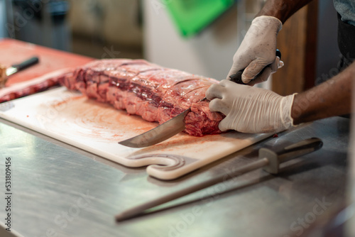 A close up of a large prime rib roast is sitting on a brown plastic cutting board. The meat has some marbling and multiple ribs. The chef is using a long knife to cut steaks from the fresh meat.