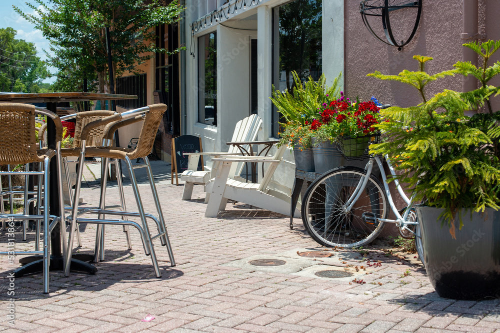 A courtyard garden outside a bakery shop with multiple wooden chairs, tables and bar stools. There's a white bicycle parked among small trees and flower pots. The store has large glass windows. 