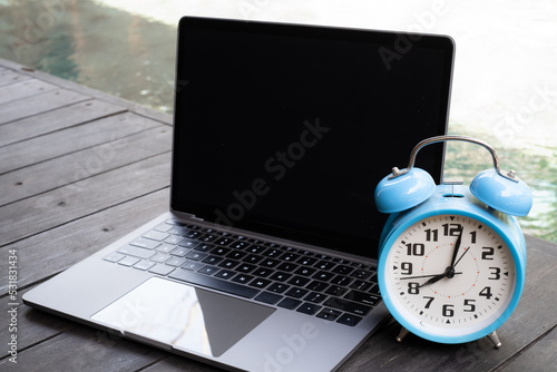 Blue vintage alarm clock on notebook near a swimming pool in morning of summer. Summer travel concept. Vacation and holiday concept. Freelance concept.