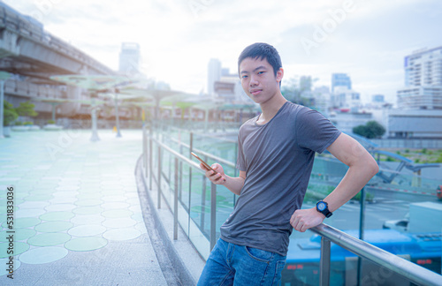 A young Asian boy, tall, smart, handsome, wears a t-shirt, jeans, stands and plays with his smart phone in the middle of the city in the morning when the sun is shining. with the use of technology