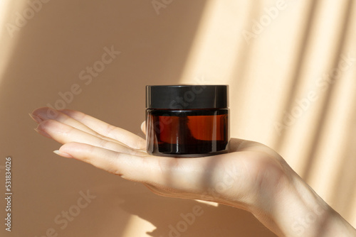 Fotobehang On the palm of woman's hand is jar of moisturizing cream made of amber glass on brown background