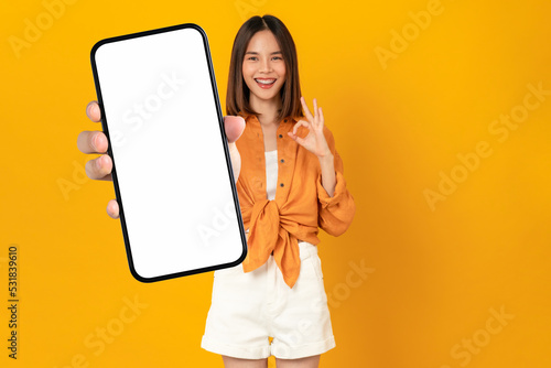 Beautiful Asian woman holding smartphone mockup of blank screen and shows ok sign on orange background.