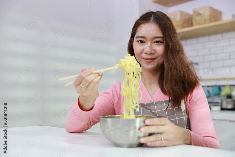 Portrait of young happy asian currly hair woman sitting on table and eating tasty and instant noodle for lunch in the kitchen with chopsticks and brick wall. Unhealthy and cheap food lifestyle concept Portrait of young happy asian currly hair woman sitting on table and eating tasty and instant noodle for lunch in the kitchen with chopsticks and brick wall. Unhealthy and cheap food lifestyle concept