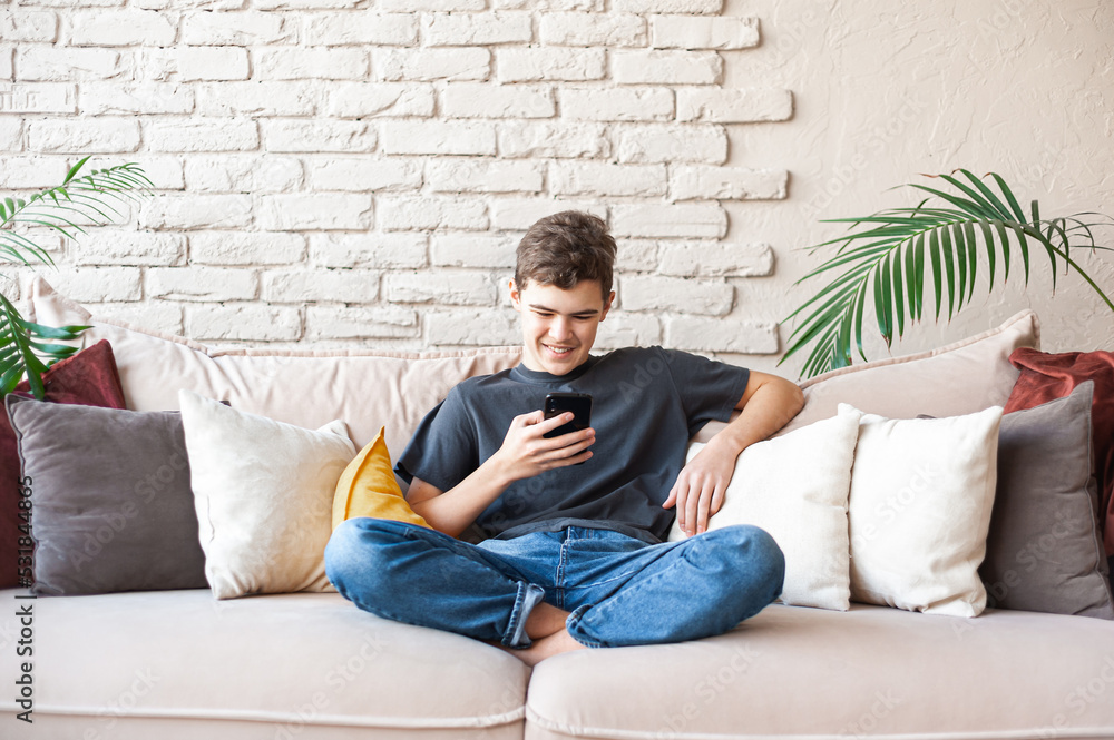teenage boy in glasses, a gray T-shirt and jeans sits in a relaxed position on the couch with a smartphone in his hands. Communicate with the use of wireless technologies. Online meetings with peers