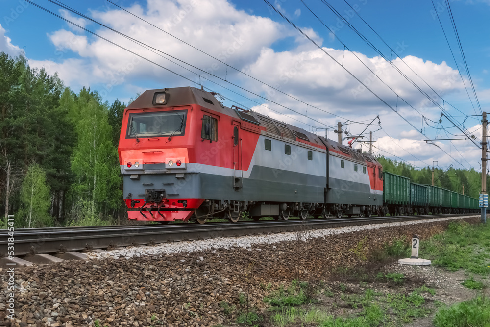 A red electric locomotive with a freight train moves along the railroad tracks. A red electric locomotive with a freight train moves along the railroad tracks.