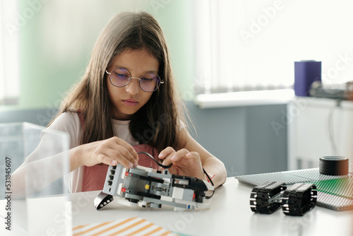 Cute serious schoolgirl creating robot or model of machine during lesson while sitting by desk and connecting parts of new item