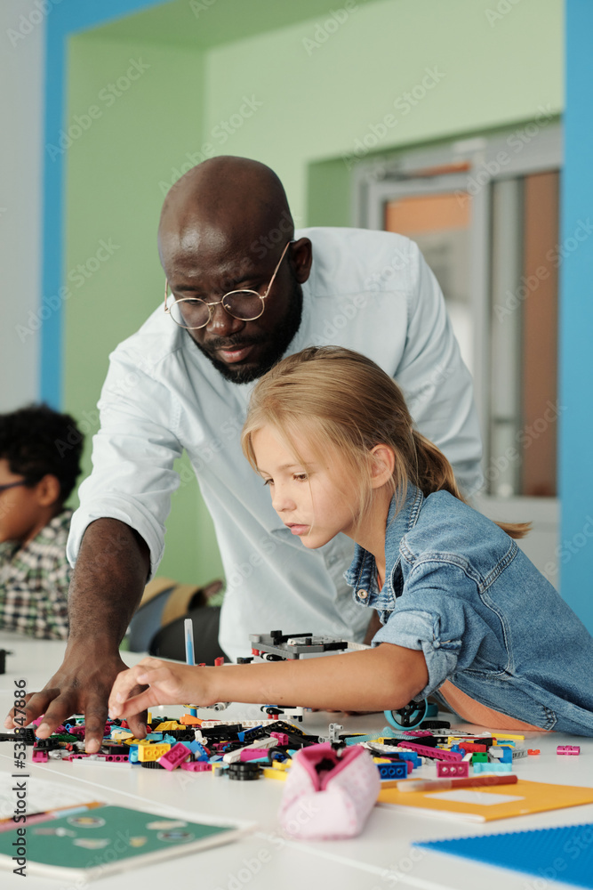 Cute schoolgirl choosing details for construction of robot with help of teacher standing next to her and bending over workplace Cute schoolgirl choosing details for construction of robot with help of teacher standing next to her and bending over workplace