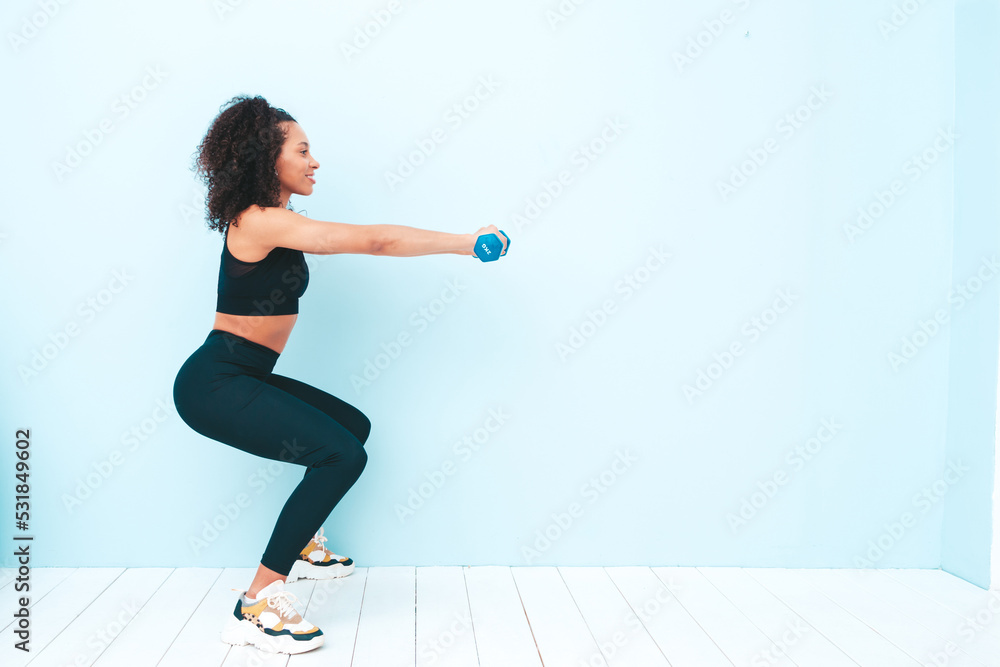 Fitness smiling black woman in sports clothing with afro curls hairstyle.She wearing sportswear. Young beautiful model doing squats.Female holding dumbbells in studio near light blue wall