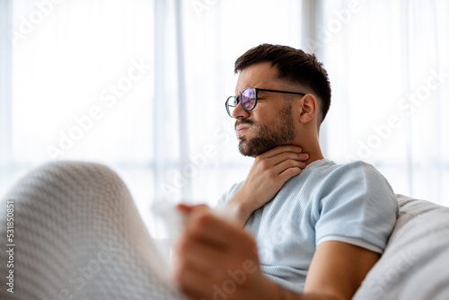 Sick man lying on the couch with high temperature and holding his throat with hand. photo