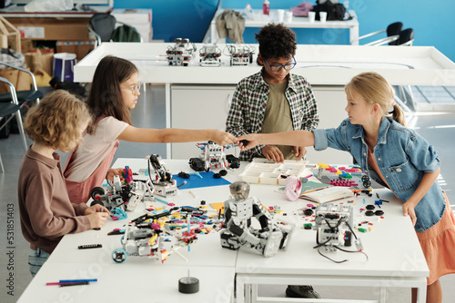 Group of youthful intercultural schoolkids constructing new robots at lesson while helpful schoolgirl passing detail to classmate photo