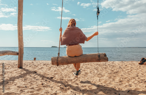 A beautiful woman is swinging on a swing overlooking the sea. Autumn at sea. Sand, water, swing. Rest by the water.
