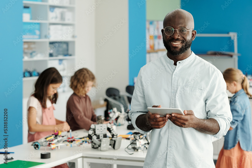 Happy young successful robotics teacher with tablet looking at camera against group of schoolkids constructing robots at lesson Happy young successful robotics teacher with tablet looking at camera against group of schoolkids constructing robots at lesson
