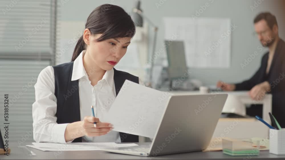 Waist up slowmo of serious female Asian engineer in formalwear doing paperwork while sitting by computer in contemporary office working there with male colleague Waist up slowmo of serious female Asian engineer in formalwear doing paperwork while sitting by computer in contemporary office working there with male colleague