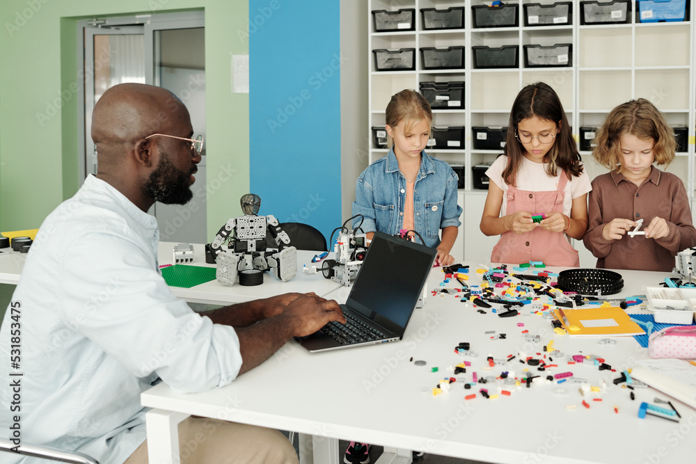 Fototapeta premium Young teacher of robotics with laptop sitting by table in front of group of schoolkids connecting parts of constructor to create robots