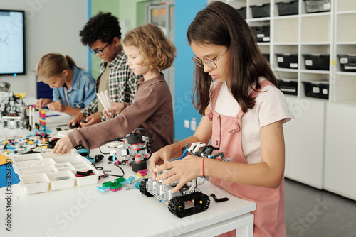 Row of four intercultural learners of elementary school constructing new toy robots while standing by table with details in classroom