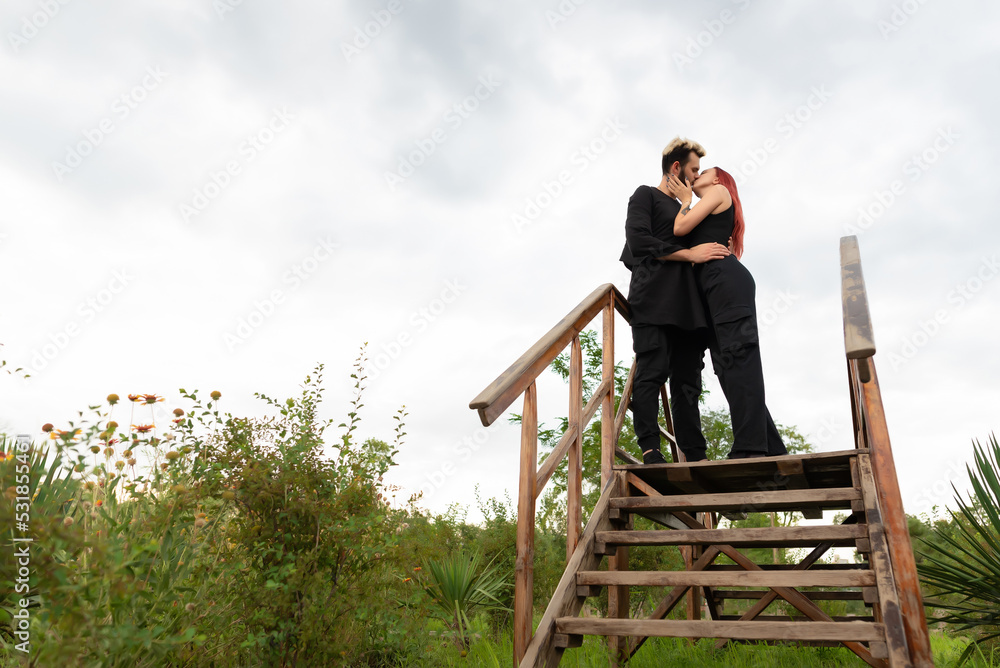 Stylish couple in love in black clothes walks in the park and hugs. A lovely couple of hipsters are standing on a wooden bridge and kissing. Youth, love and lifestyle concept