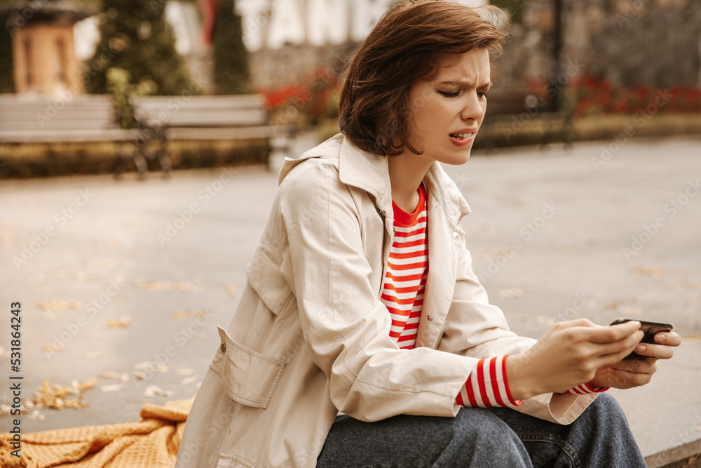 Young caucasian woman looking at smartphone upset by bad reading news on phone while sitting street. Brunette girl with bob haircut wears coat, jacket and jeans. Leisure time concept Young caucasian woman looking at smartphone upset by bad reading news on phone while sitting street. Brunette girl with bob haircut wears coat, jacket and jeans. Leisure time concept