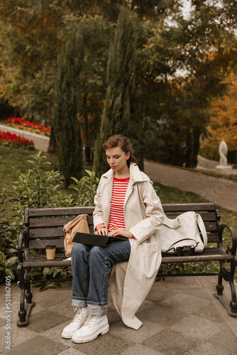 Full length concentrated young caucasian girl sitting on bench using tablet pc outside. Brunette wears jacket, jeans, coat and boots. Social media addiction concept