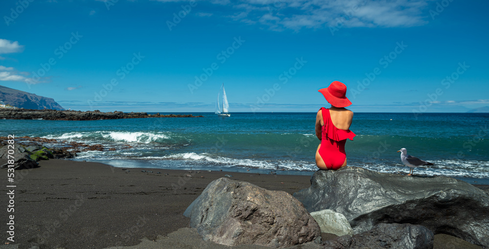 Woman in red swimming suit sitting on the rock in the black ocean sandy beach. Calm ocean water and pure blue sky at the background. Woman in red swimming suit sitting on the rock in the black ocean sandy beach. Calm ocean water and pure blue sky at the background.