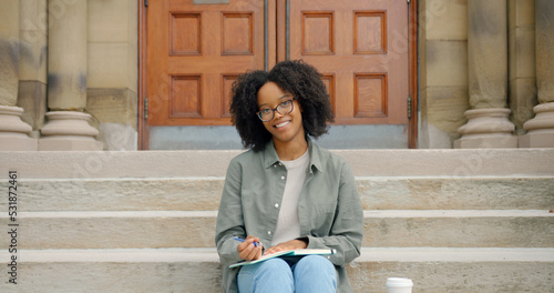 African charming young lady with curly hair and glasses, smiling toothily seats on the stairs outside and writing in her notepad © South House Studio