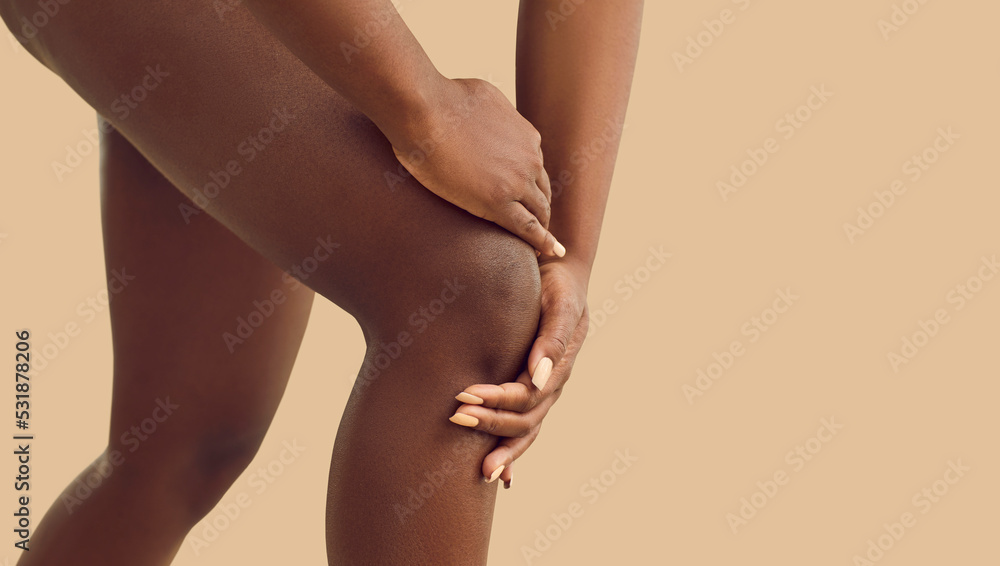 Young black lady whose knee is hurting is holding her hand on injured knee cap area. Cropped shot of human legs isolated on beige color background. Injury, rheumatoid arthritis, feeling pain concept