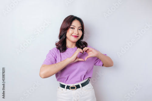 An attractive young Asian woman wearing a lilac purple t-shirt feels happy and a romantic shapes heart gesture expresses tender feelings