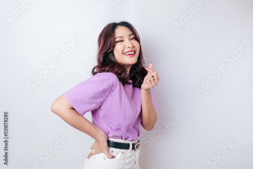 An attractive young Asian woman wearing a lilac purple t-shirt feels happy and a romantic shapes heart gesture expresses tender feelings
