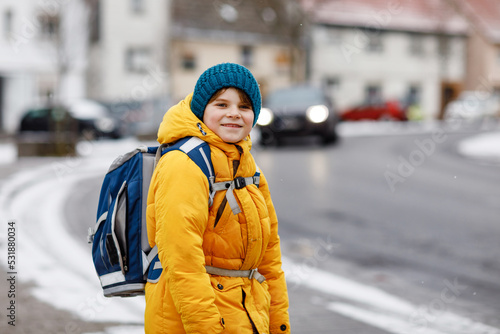 Little school kid boy of elementary class walking to school during snowfall. Happy child having fun and playing with first snow. Student with in yellow jacket and backpack in colorful winter clothes.
