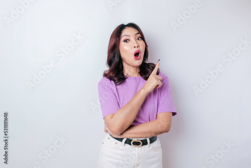 Shocked Asian woman wearing lilac purple t-shirt pointing at the copy space upside her, isolated by white background
