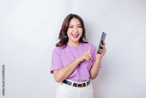 A portrait of a happy Asian woman is smiling and holding her smartphone wearing a lilac purple t-shirt isolated by a white background