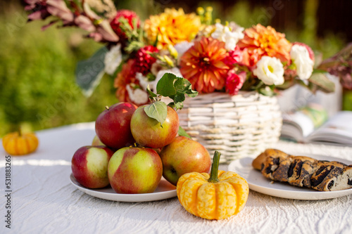 Ripe apples are lying on the table near a small pumpkin and seasonal flowers