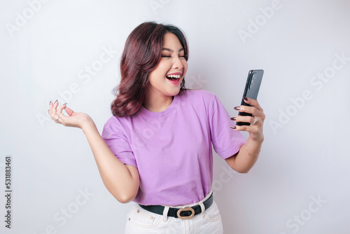 A portrait of a happy Asian woman is smiling and holding her smartphone wearing a lilac purple t-shirt isolated by a white background