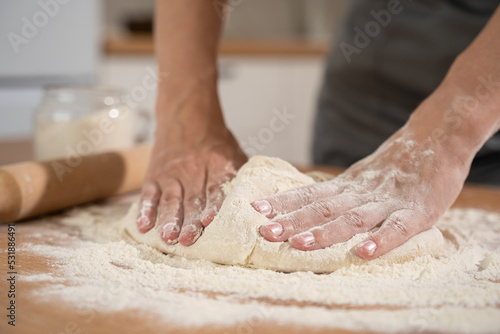 A young woman in a gray apron throws a ball of dough on the wooden surface of the kitchen table with a pile of flour. 