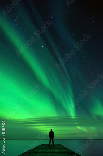 Man standing on pier and watching Northern Lights dancing across night sky above lake landscape.