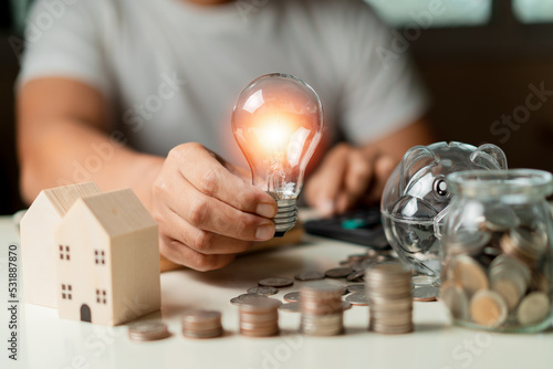 Businessman using calculator to calculate and holding a light bulb and Stacking coins, piggy, wooden house on table. Saving money,accounting, investment, budgeting, and financial planning concept.