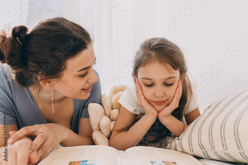 Close-up image of elder sister spending time with her younger one reading her book lying on bed  looking at girl with love and care  kid listening attentively leaning chin on hands