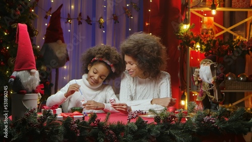 African American mom and little daughter decorating New Year's figures with paints. Woman and girl sitting near Christmas tree. Happy family portrait. New Year celebration concept.