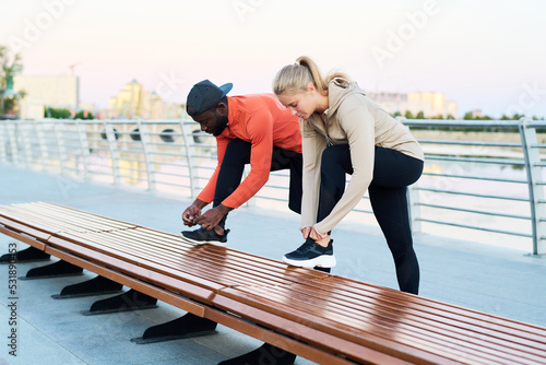 Young intercultural man and woman in activewear tying shoelaces on sneakers while preparing for outdoor sports training