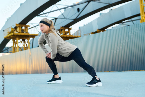 Young fit blond woman in activewear doing physical exercise for leg stretching while standing on asphalt against modern architecture