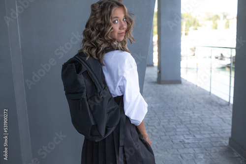 Beautiful fashionable school student, dressed in a white blouse and black skirt holds a black backpack with lessons and smiles