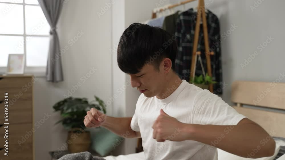 tired asian male rising early in the morning is sitting on bed and warming up his body by stretching arms in a modern bedroom at home tired asian male rising early in the morning is sitting on bed and warming up his body by stretching arms in a modern bedroom at home