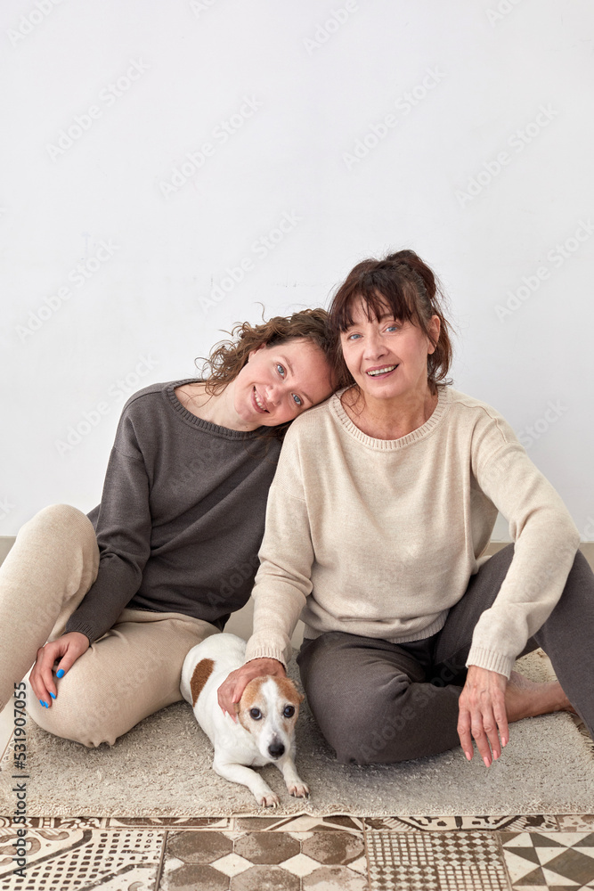 Cheerful adult daughter and middle aged mother sitting on floor with cute dog at home Cheerful adult daughter and middle aged mother sitting on floor with cute dog at home