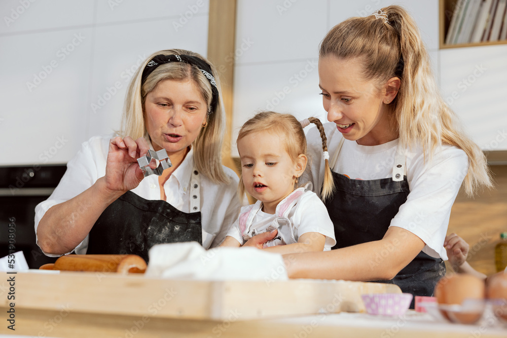 Close up shot granny and mother kneading dough with preschooler offspring in kitchen standing at table. Pretty girl having fun with family granny holding baking molds in arm. Close up shot granny and mother kneading dough with preschooler offspring in kitchen standing at table. Pretty girl having fun with family granny holding baking molds in arm.