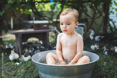 Little funny boy is bathing in a big metal trough. A child bathes in a trough surrounded by ducks. Village fun. Rural life. Happy childhood. The child is bathing in the garden.