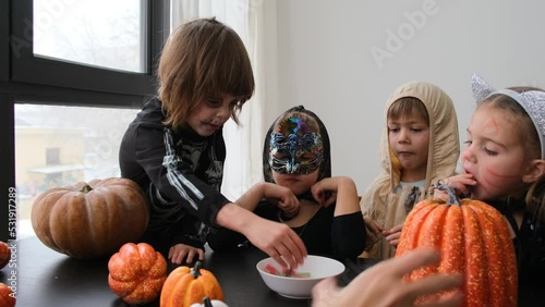 Long haired junior schoolgirls and preschooler brother in Halloween costumes eat treats sitting at table decorated with pumpkins at home
