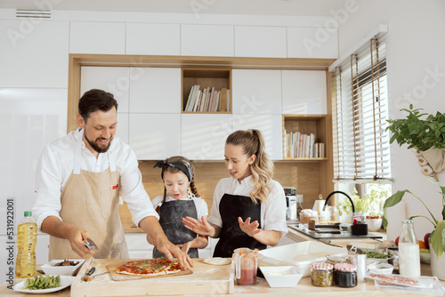 Curious kid child daughter looking at homemade pizza with mother sniffing flavour handsome father cutting pizza with pizza cutter smiling. Delicious tasty dish on wooden table.