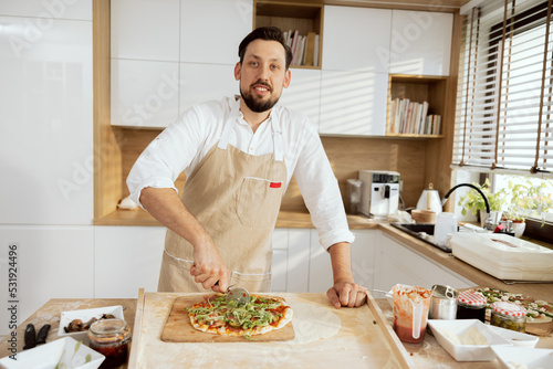 Handsome delighted man in aprons cutting with pizza cutter domestic pizza on wooden surface in beautiful modern light kitchen with large window.