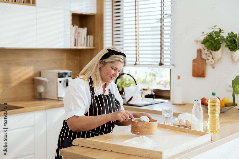 Sieving flour on wooden surface wearing apron smiling. Delighted woman spends time in modern light kitchen. Baking process preparing surprise f Sieving flour on wooden surface wearing apron smiling. Delighted woman spends time in modern light kitchen. Baking process preparing surprise f