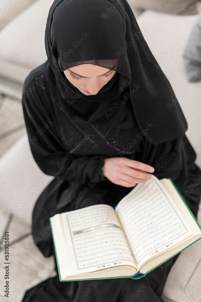Young traditional muslim woman reading Quran on the sofa before iftar dinner during a ramadan feast at home Young traditional muslim woman reading Quran on the sofa before iftar dinner during a ramadan feast at home