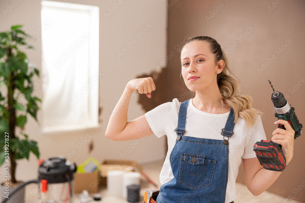 a girl dressed in denim style, holds a drill in her left hand, and shows her biceps with her right hand. In the background there are many things to repair.